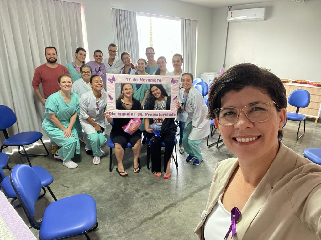 Grupo de profissionais de saúde e outras pessoas reunidas em uma sala com cadeiras azuis, sorrindo para a câmera. Em primeiro plano, uma mulher de óculos e cabelo curto tira a selfie, vestindo blazer claro com laço roxo preso na lapela. Ao fundo, o grupo segura uma moldura decorada com borboletas e a frase 'Dia Mundial da Prematuridade - 17 de Novembro'. Alguns usam uniformes hospitalares em tons de verde e branco, enquanto outros estão com roupas casuais.