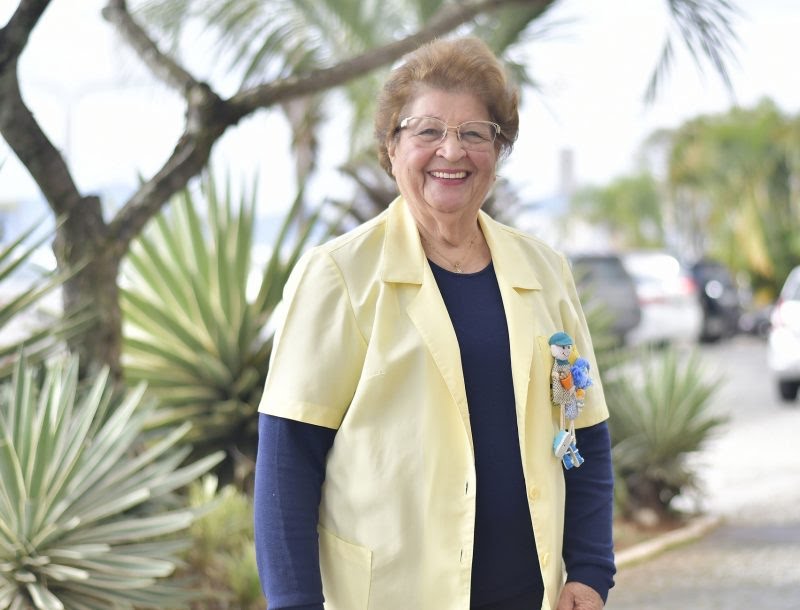 Senhora idosa sorridente usando óculos e jaleco amarelo sobre blusa azul escura, posando ao ar livre em calçada arborizada com palmeiras e carros estacionados ao fundo. Ela tem um broche decorativo colorido preso ao jaleco.