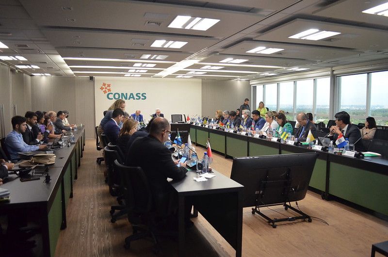 Reunião formal com dezenas de pessoas sentadas ao redor de uma grande mesa em formato de U, dentro de uma sala moderna com janelas amplas. No fundo da sala, a palavra "CONASS" está escrita na parede. As mesas possuem placas de identificação e bandeiras de diversos estados brasileiros, sugerindo uma reunião oficial de representantes estaduais da saúde.