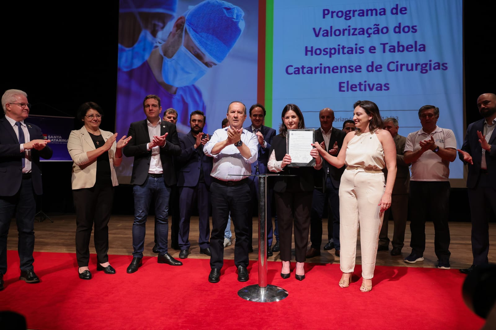A imagem mostra um grupo de autoridades e representantes reunidos em um palco para um evento oficial.  No centro, duas mulheres seguram e apresentam um documento oficial; uma delas veste roupa clara e a outra, roupa preta.  Ao lado delas, um homem de camisa social branca aplaude, assim como outras pessoas posicionadas em semicírculo atrás.  O palco tem um tapete vermelho e um púlpito metálico no centro.  No telão ao fundo, lê-se: "Programa de Valorização dos Hospitais e Tabela Catarinense de Cirurgias Eletivas".  Ao lado do texto, há uma imagem de um profissional de saúde com máscara e touca cirúrgica.  Trata-se de um momento solene, possivelmente de assinatura ou anúncio oficial ligado à saúde pública do estado de Santa Catarina.