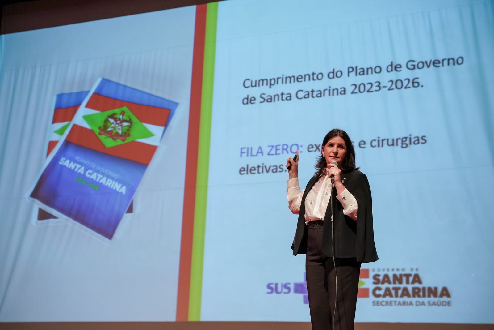 A imagem mostra uma mulher apresentando em um auditório, em frente a um telão com projeção.  Ela segura um microfone na mão esquerda e um controle remoto ou apontador na mão direita.  Na tela, aparecem dois livros com a capa da bandeira de Santa Catarina e o título “Plano de Governo Santa Catarina 2023-2026”.  Ao lado, está escrito: "Cumprimento do Plano de Governo de Santa Catarina 2023-2026. FILA ZERO: exames e cirurgias eletivas".  Na parte inferior direita da projeção, está o logotipo do Governo de Santa Catarina – Secretaria da Saúde junto ao símbolo do SUS.  O fundo do slide tem faixas verticais nas cores da bandeira catarinense (vermelho, amarelo e verde).