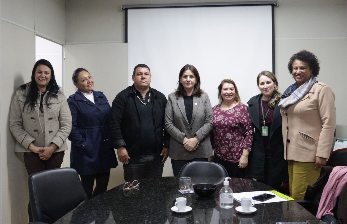 Sete pessoas posam para foto em sala de reuniões, com mesa redonda à frente e tela de projeção ao fundo. Entre elas, representantes indígenas e autoridades públicas. Reunião formal com foco em políticas de saúde para povos indígenas.
