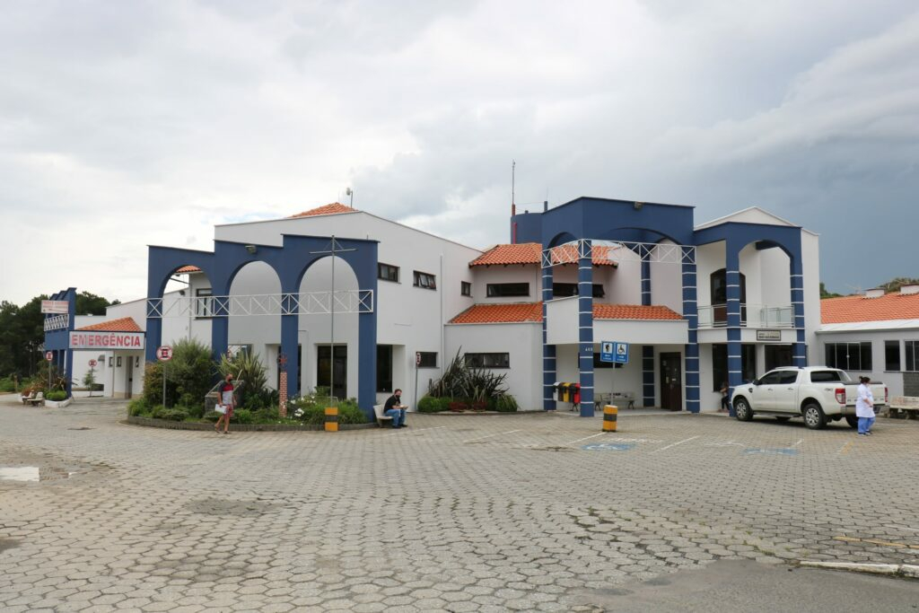 Fachada de um hospital com arquitetura moderna e detalhes em azul e branco. À esquerda, placa com a palavra "Emergência". No pátio frontal, pessoas circulam e há veículos estacionados, incluindo uma caminhonete branca. O céu está nublado.