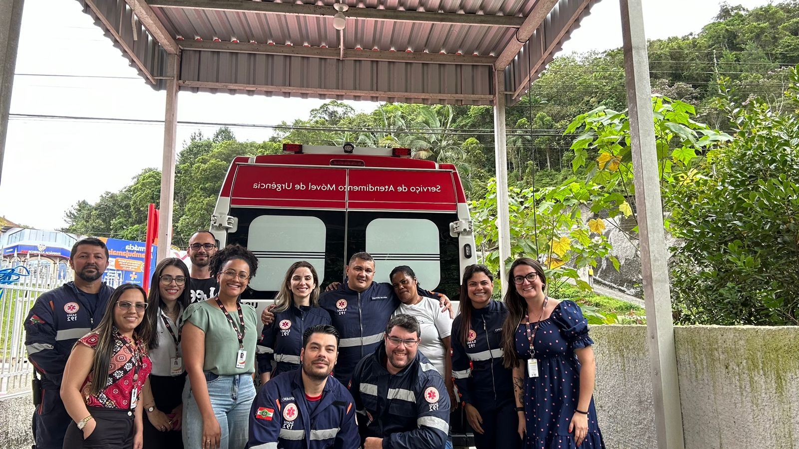 A imagem mostra um grupo de pessoas posando juntas em frente a uma ambulância do SAMU (Serviço de Atendimento Móvel de Urgência).  Alguns integrantes usam uniformes azuis característicos do serviço, com faixas refletivas e o número 192, enquanto outros estão vestidos de forma casual ou com crachás de identificação, sugerindo que possam ser profissionais de saúde ou apoio administrativo.  O cenário é uma área coberta, com vegetação ao fundo e parte de um portão lateral visível à esquerda. O clima da foto é de união e cooperação, transmitindo a ideia de trabalho em equipe e compromisso com o atendimento de urgência e emergência.