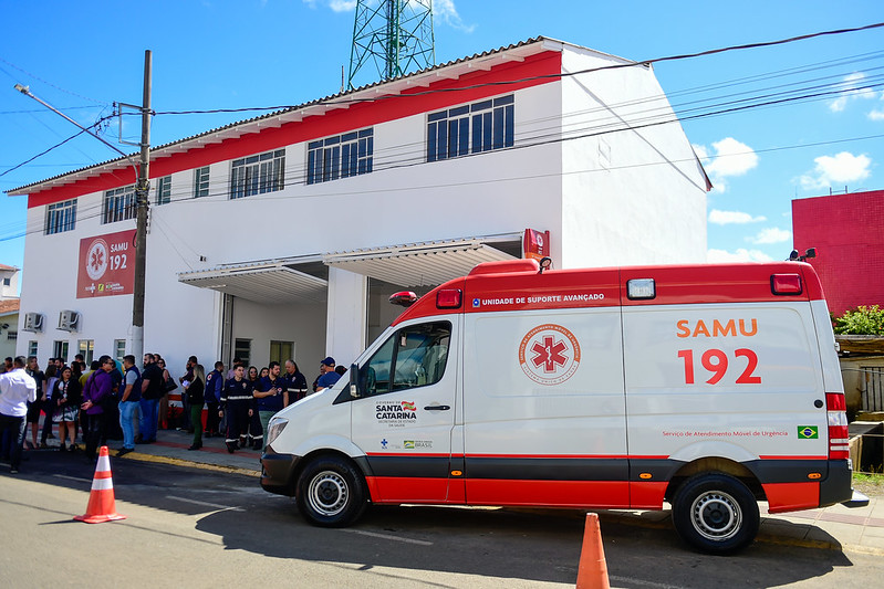 Imagem da base do SAMU em São Joaquim, Santa Catarina. Em frente ao prédio branco com detalhes em vermelho e a logomarca do SAMU 192, há uma ambulância de Unidade de Suporte Avançado estacionada. Ao lado da ambulância, um grupo de pessoas está reunido, provavelmente para uma cerimônia ou inauguração. No veículo e no prédio, estão visíveis os logotipos do Governo de Santa Catarina e do Ministério da Saúde. O céu está azul e o clima ensolarado.