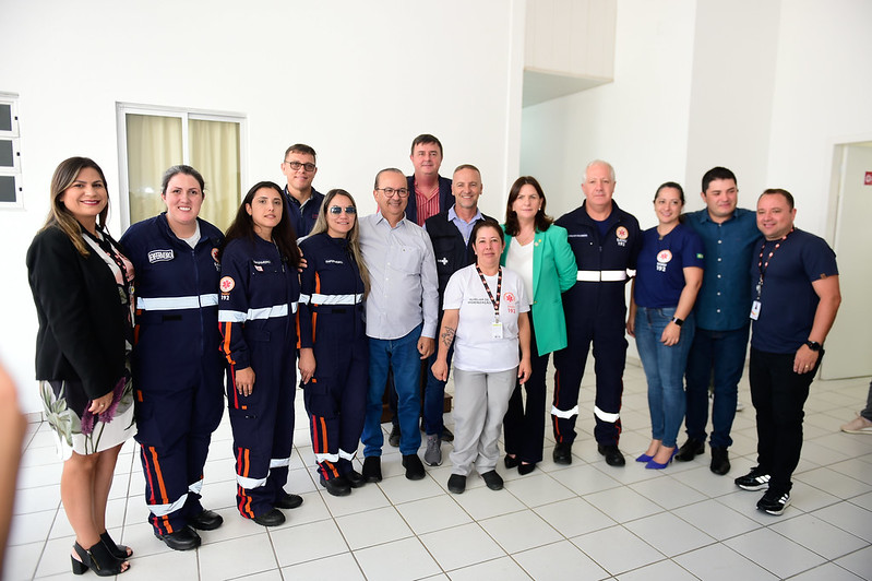 Grupo de profissionais do SAMU e autoridades posando para uma foto em ambiente interno com paredes e piso brancos. Ao todo, são 16 pessoas, entre homens e mulheres, alguns trajando uniformes do SAMU (macacão azul com faixas refletivas e detalhes em laranja), outros com roupas casuais ou formais. Todos estão sorrindo, demonstrando um momento de confraternização ou celebração institucional.