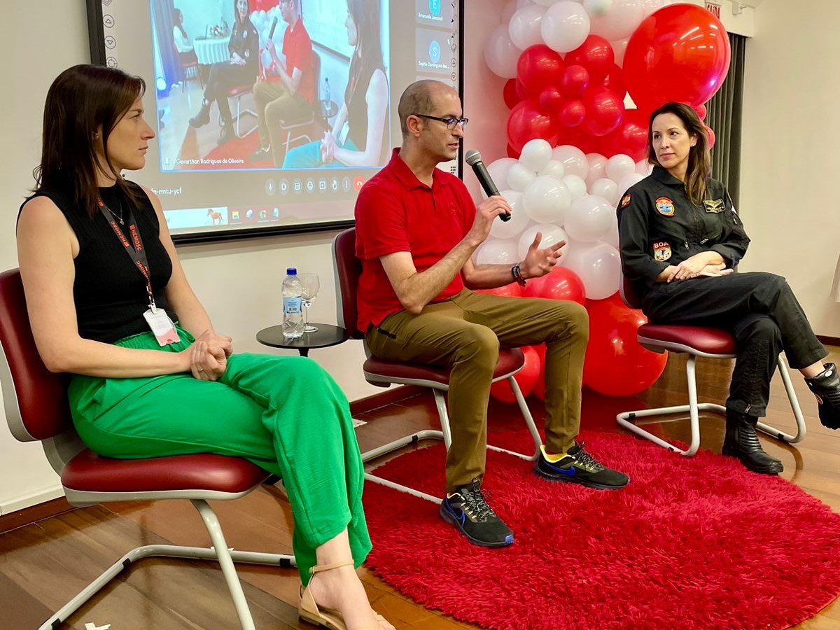 Três palestrantes sentados em cadeiras durante evento sobre doação de sangue, com balões vermelhos e brancos ao fundo e uma apresentação projetada na tela.