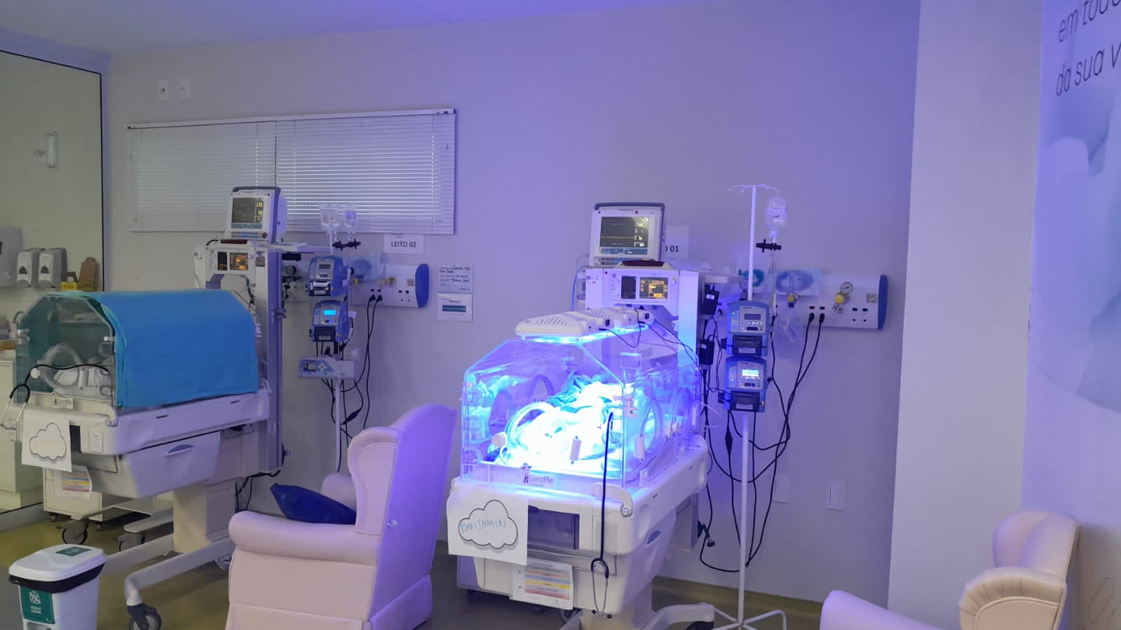 Unidade de Terapia Intensiva Neonatal com incubadoras modernas e equipamentos de suporte à vida. Um dos berços exibe luz azul, indicando o uso de fototerapia em um recém-nascido. Ao fundo, monitores, bombas de infusão e outros aparelhos médicos estão conectados, destacando o ambiente altamente controlado e especializado.