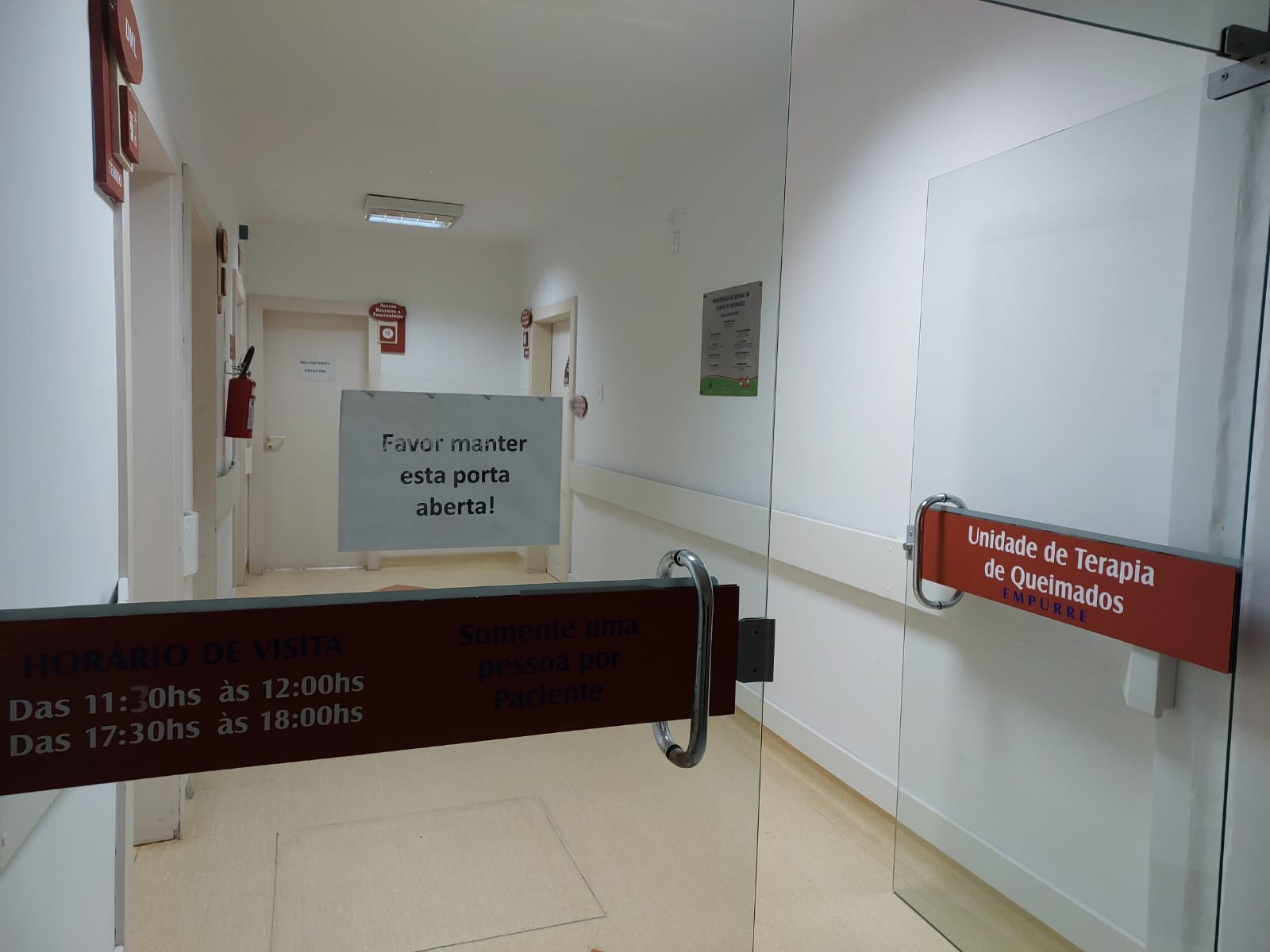 A imagem mostra a entrada de um corredor hospitalar, vista através de portas de vidro. No vidro à direita, há uma placa vermelha com os dizeres “Unidade de Terapia de Queimados – Empurre”. No vidro à esquerda, outra placa vermelha informa o horário de visitas:  Das 11:30 às 12:00  Das 17:30 às 18:00 E acrescenta: “Somente uma pessoa por paciente”.  Ao fundo, fixado na parede, há um aviso em papel escrito: "Favor manter esta porta aberta!". O ambiente é bem iluminado, com paredes brancas, piso claro e extintores de incêndio posicionados nas paredes.
