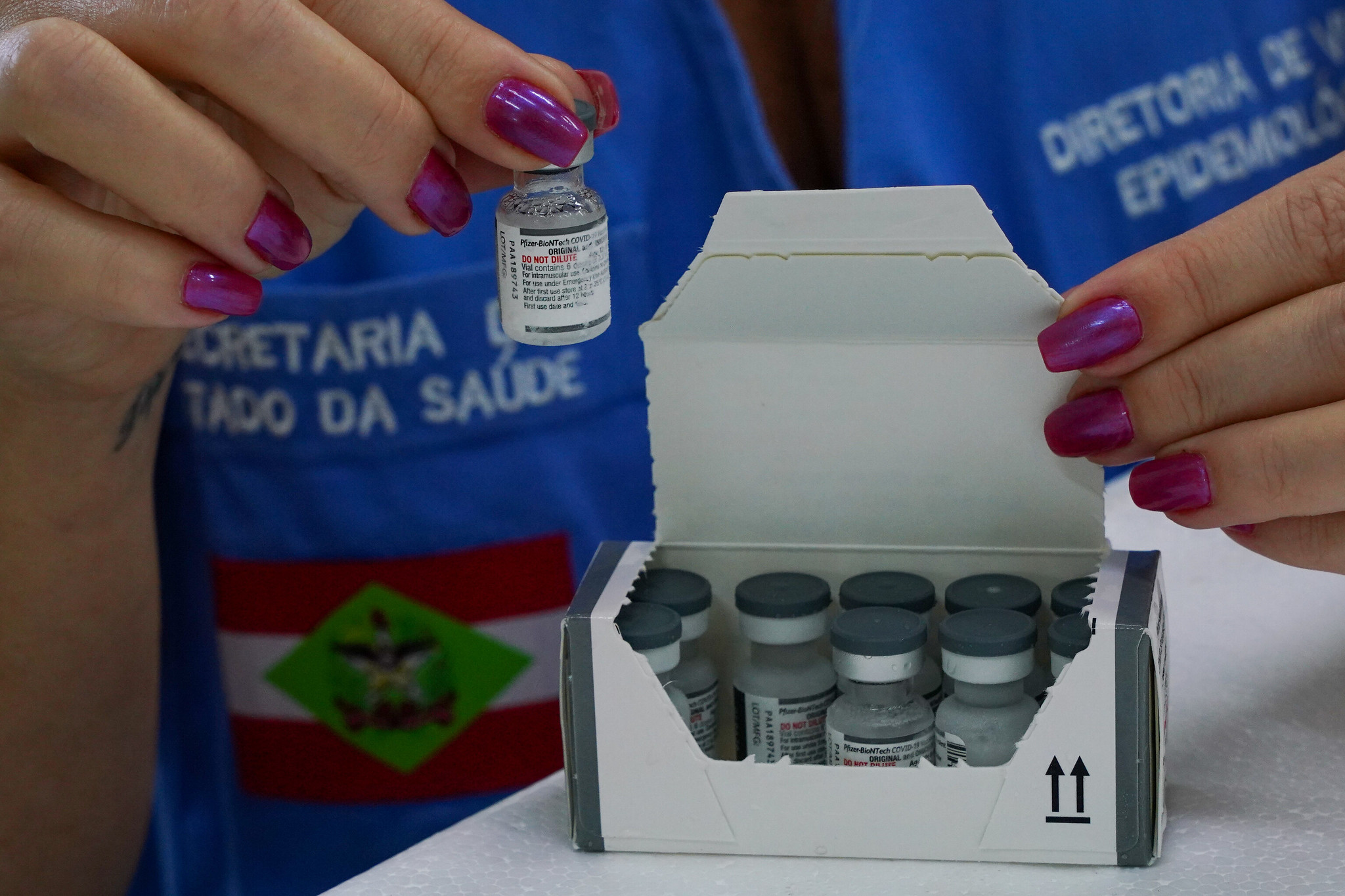 Profissional da saúde com unhas pintadas de roxo segura um frasco de vacina da Pfizer-BioNTech contra a COVID-19 acima de uma caixa com vários frascos similares. A pessoa veste colete azul com os dizeres "Secretaria de Estado da Saúde" e "Diretoria de Vigilância Epidemiológica", além da bandeira do estado de Santa Catarina.