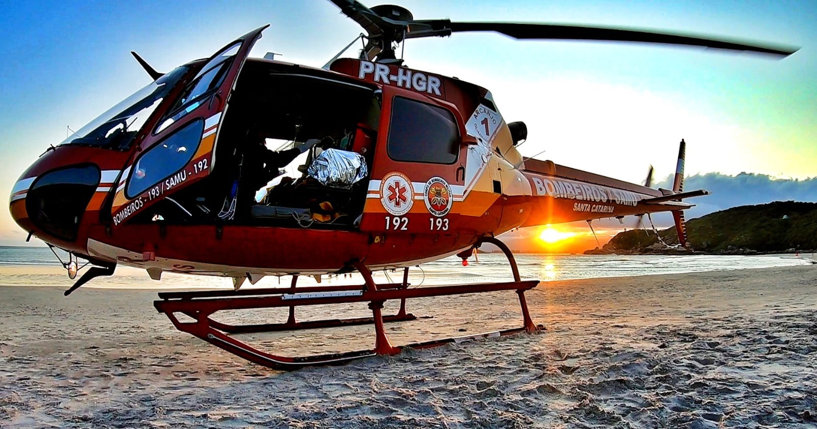 Helicóptero de resgate do Corpo de Bombeiros Militar de Santa Catarina estacionado em uma praia ao pôr do sol. A aeronave vermelha exibe os números de emergência 192 (SAMU) e 193 (Bombeiros), com as portas abertas e parte do interior visível. Ao fundo, o sol se põe atrás de colinas, refletindo sobre o mar.