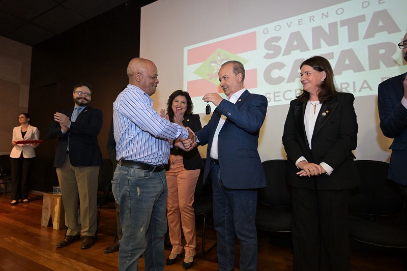 Homem de camisa listrada e jeans recebe simbolicamente uma chave de outro homem de terno azul durante uma cerimônia. Eles apertam as mãos enquanto outras autoridades, incluindo mulheres e homens de terno, observam e aplaudem. Ao fundo, o telão exibe o logotipo do Governo de Santa Catarina e da Secretaria da Saúde.