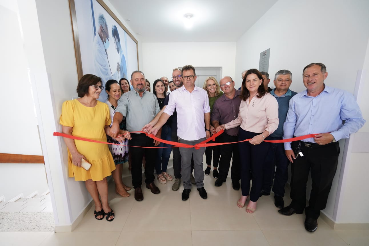 Grupo de pessoas participando de uma cerimônia de inauguração, posando para foto enquanto seguram uma fita vermelha prestes a ser cortada. A cena ocorre em um ambiente interno, iluminado, com paredes claras e piso de porcelanato. Ao fundo, um quadro grande com imagem de profissionais da saúde em atendimento. O clima é de celebração institucional.