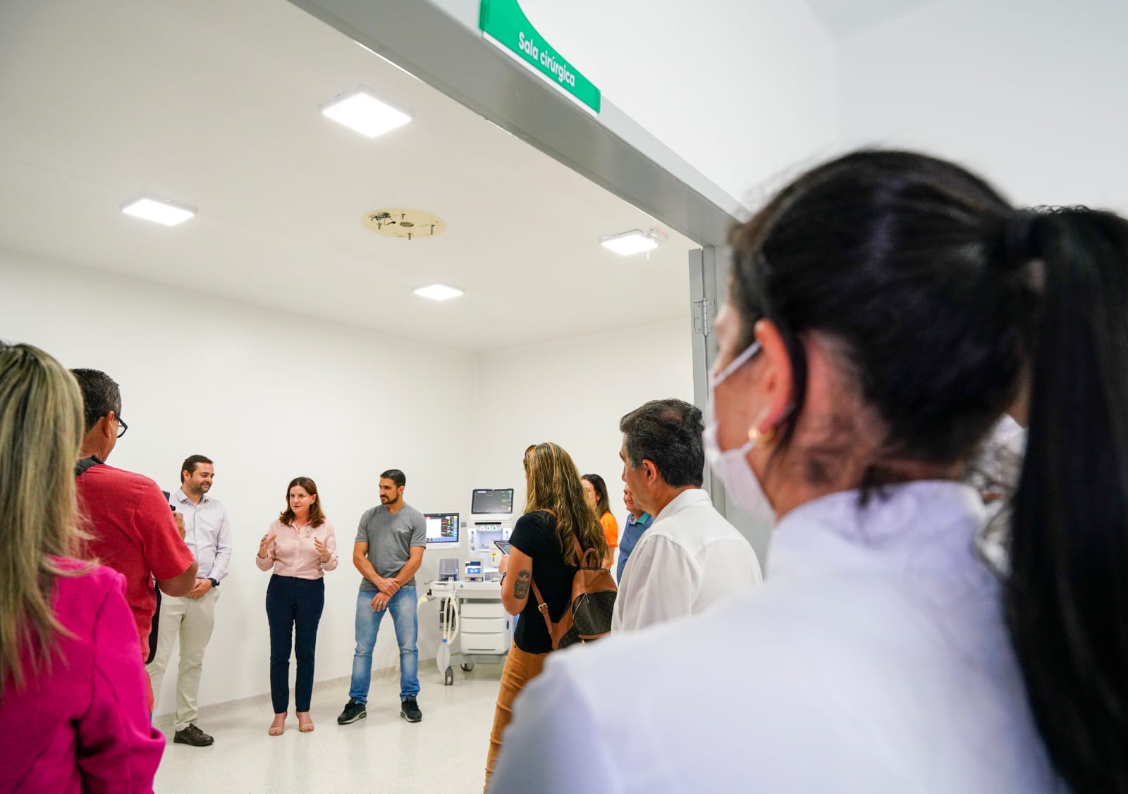 Pessoas reunidas na inauguração de uma sala cirúrgica, observando três profissionais (dois homens e uma mulher) que falam próximos a equipamentos hospitalares modernos. O ambiente é bem iluminado, com paredes e piso claros, e uma placa verde com o texto “Sala cirúrgica” identifica o local. A maioria dos presentes usa roupas sociais ou jalecos, e alguns utilizam máscaras de proteção.