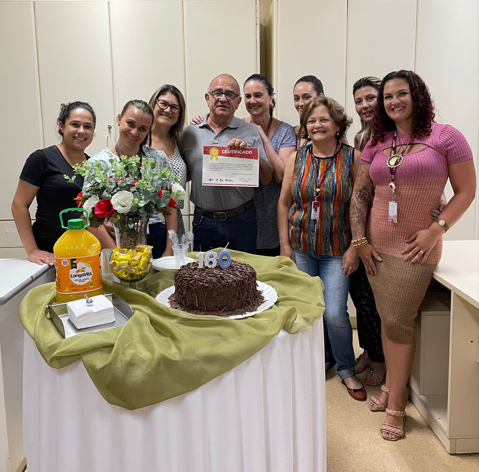 Grupo de nove pessoas posa sorridente ao redor de uma mesa decorada com toalha branca e tecido verde. Sobre a mesa, há um bolo de chocolate com velas formando o número 160, um vaso com flores artificiais, suco de laranja e balas. Ao centro, um homem segura um certificado, aparentando estar sendo homenageado. Todos demonstram alegria em um ambiente de confraternização.