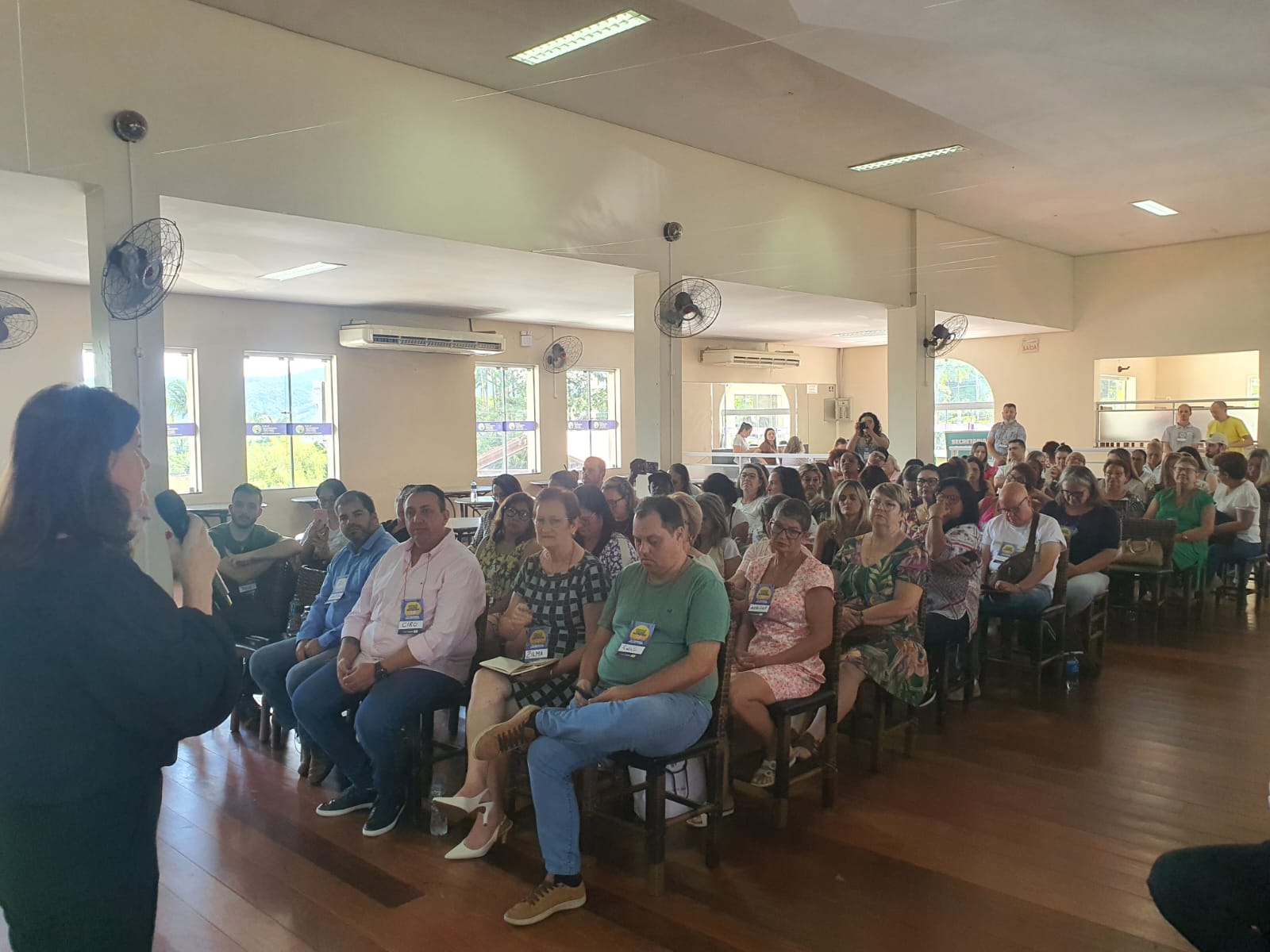 Mulher fala ao microfone diante de um auditório cheio, onde diversas pessoas estão sentadas em cadeiras de madeira, prestando atenção à palestra. O evento ocorre em um ambiente interno, iluminado por luz natural que entra pelas janelas. Algumas pessoas usam crachás de identificação. O clima é de atenção e engajamento.