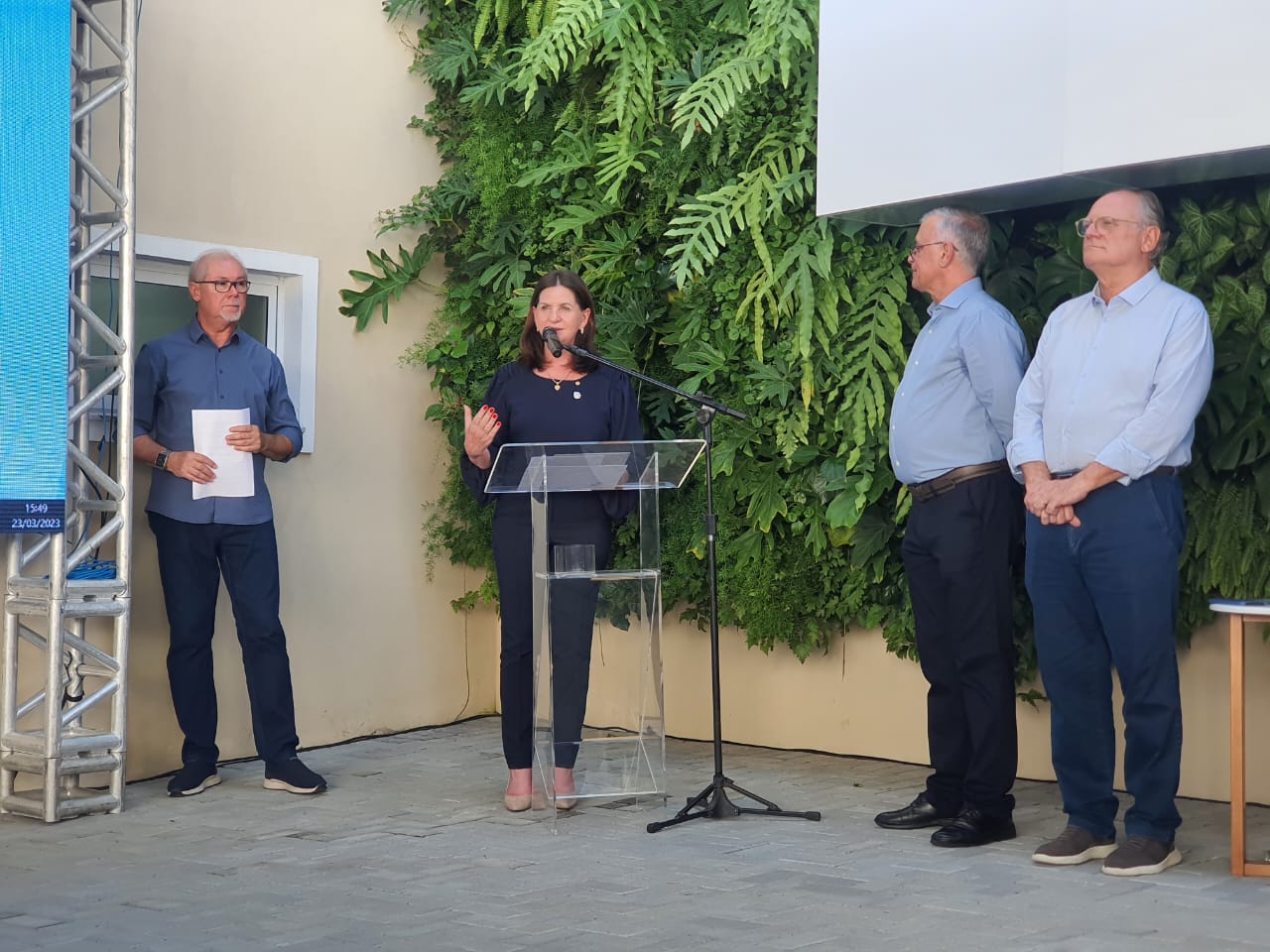 Mulher fala ao microfone em um púlpito de acrílico durante um evento ao ar livre, com três homens ao fundo, todos vestidos com roupas sociais. Ao lado esquerdo da imagem, há uma estrutura metálica com uma tela digital azul, e ao fundo, uma parede verde com plantas naturais. A cena acontece durante o dia, em ambiente iluminado. No canto inferior esquerdo, vê-se um relógio digital com a data “23/03/2023”.