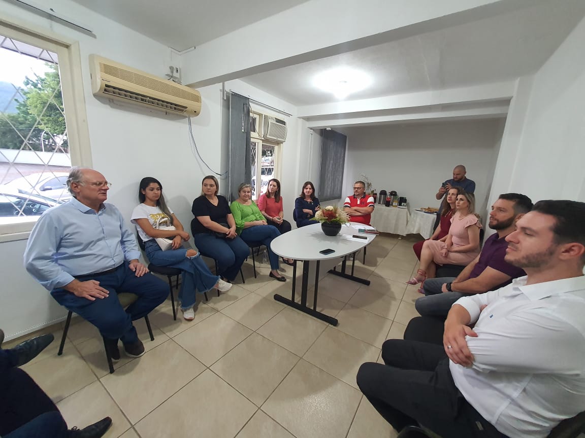 Reunião em sala fechada com pessoas sentadas em círculo, demonstrando atenção e participação. Há uma mesa central com um vaso de flores e, ao fundo, uma mesa de café com garrafas térmicas e lanches. As pessoas estão vestidas de maneira informal e parecem estar envolvidas em uma conversa ou discussão.