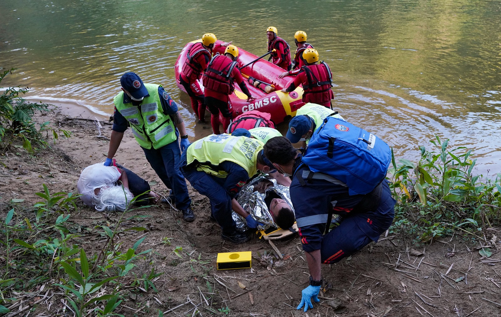 Equipe de resgate com coletes e capacetes realiza o salvamento de duas vítimas às margens de um rio. Uma balsa inflável vermelha com o logo do Corpo de Bombeiros Militar de Santa Catarina (CBMSC) está parcialmente na água, enquanto socorristas atendem uma pessoa enrolada em manta térmica e outra deitada no chão. A vegetação ribeirinha envolve a cena.