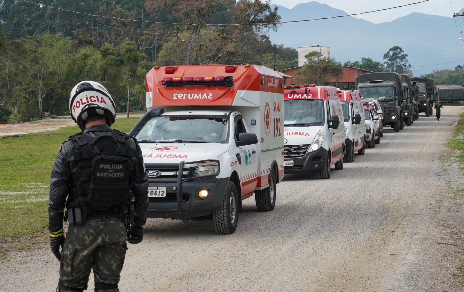 Policial do Exército com uniforme tático e capacete branco observa comboio formado por ambulâncias do SAMU e veículos militares em estrada de terra. Ao fundo, há árvores, montanhas e edificações, sugerindo uma operação conjunta de apoio ou simulado de emergência.