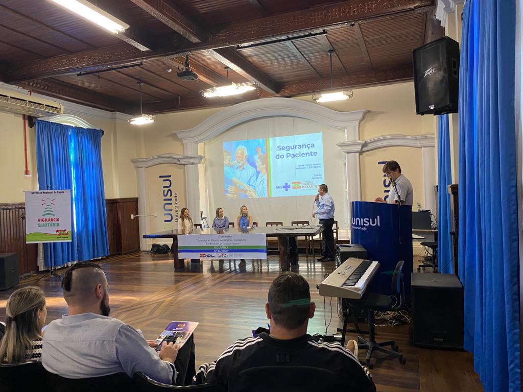 Palestra sobre Segurança do Paciente em auditório da Unisul, com painel projetado ao fundo, quatro pessoas sentadas à mesa, um palestrante ao microfone e participantes assistindo. Há uma placa da Vigilância Sanitária ao lado do palco.