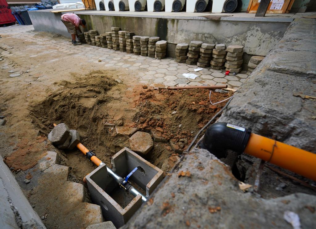 Homem trabalha em obra de instalação hidráulica externa, com tubulação laranja e caixa de inspeção de concreto parcialmente enterrada; ao fundo, vários blocos de piso intertravado empilhados aguardam reinstalação.