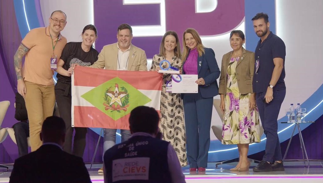 A imagem mostra um grupo de sete pessoas no palco durante um evento, posando para uma foto oficial.  No centro, duas mulheres seguram um certificado e um troféu, enquanto três pessoas à esquerda exibem a bandeira do estado de Santa Catarina. À direita, outras duas pessoas completam o grupo. O cenário de fundo apresenta iluminação roxa e branca, com letras grandes compondo a identidade visual do evento.  Todos sorriem, indicando que se trata de um momento de celebração ou premiação. Na frente do palco, é possível ver pessoas da plateia, incluindo uma com colete escrito "Rede CIEVS – Centro de Informações Estratégicas de Vigilância em Saúde".