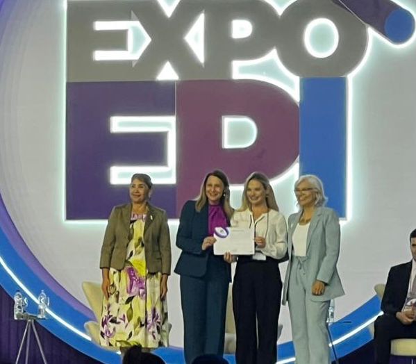 A imagem mostra quatro mulheres no palco de um evento, posando para uma foto oficial de premiação ou reconhecimento.  Ao fundo, há um grande painel iluminado com o logotipo "EXPO EPI", sugerindo que se trata de um evento relacionado à saúde pública e epidemiologia. A segunda e a terceira mulheres (da esquerda para a direita) seguram um certificado e um troféu circular, sorrindo para a câmera.  As outras duas mulheres, uma à esquerda e outra à direita, acompanham a entrega, também sorrindo. A cena transmite um clima de celebração e valorização profissional, provavelmente pelo reconhecimento de um trabalho de destaque na área.