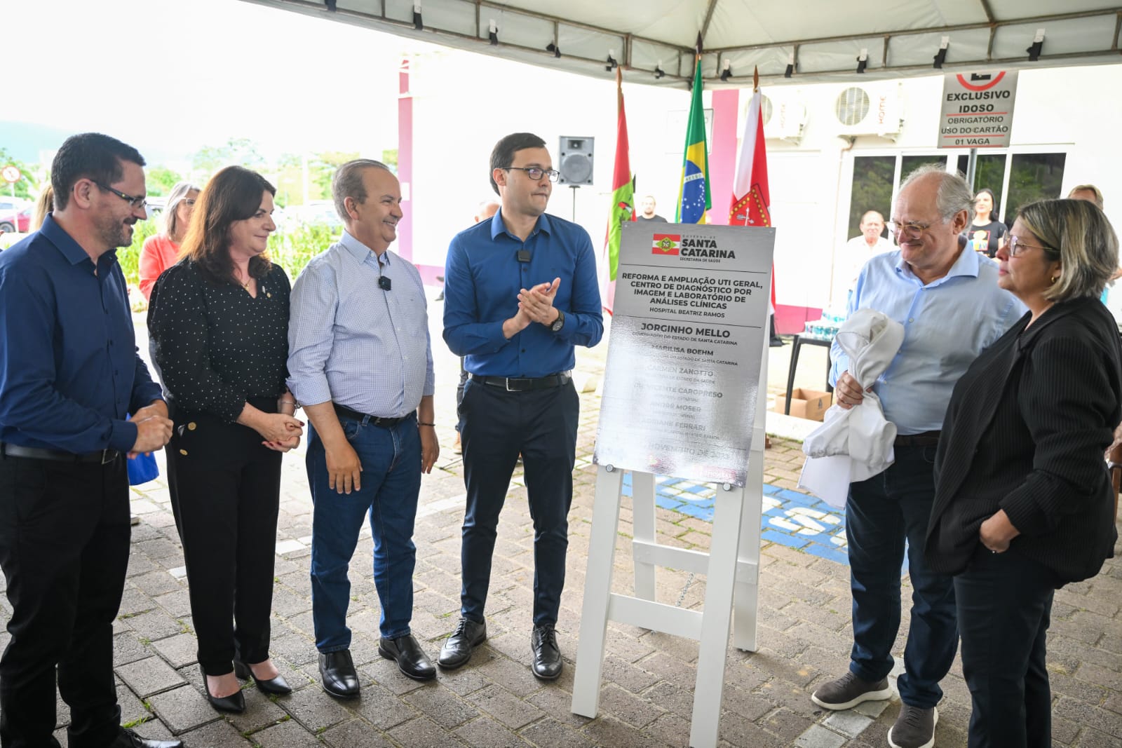 A imagem mostra uma cerimônia de inauguração ou entrega de obra pública.  Seis pessoas estão reunidas em frente a uma placa metálica com o brasão e o logotipo do Governo de Santa Catarina. A placa indica a reforma e ampliação da UTI Geral, do Centro de Diagnóstico por Imagem e do Laboratório de Análises Clínicas do Hospital Beatriz Ramos, com menção ao governador Jorginho Mello e outras autoridades.  Os participantes estão bem-vestidos, com expressão de satisfação, e alguns sorriem ou aplaudem. Ao fundo, há bandeiras de Santa Catarina e do Brasil, e um toldo que cobre a área. O clima é de evento oficial, com registro fotográfico e presença de público.