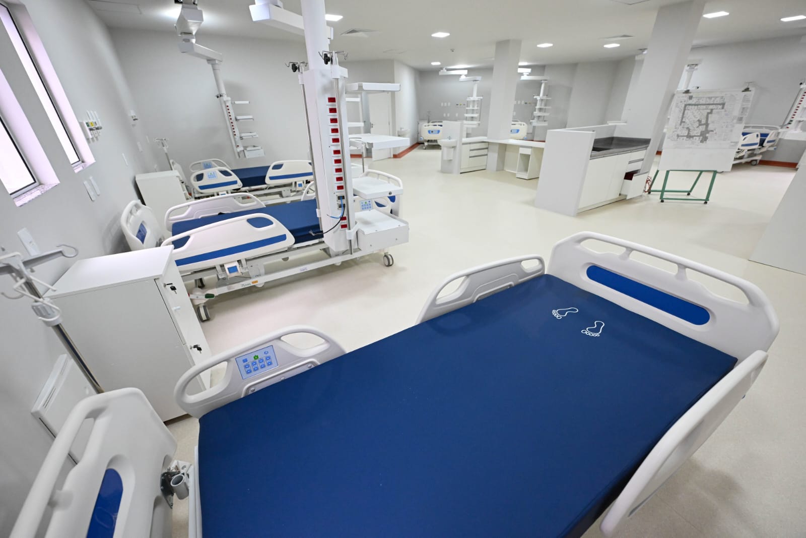 A imagem mostra uma enfermaria hospitalar nova ou recentemente reformada, equipada com diversas camas hospitalares modernas, todas com colchões azuis e cabeceiras ajustáveis.  O espaço é amplo, bem iluminado por luz artificial, com piso claro e paredes brancas. Ao fundo, é possível ver equipamentos hospitalares, armários, suportes para soro, e uma planta baixa fixada em um suporte. As camas têm painéis de controle eletrônico e estão organizadas de forma espaçosa, sugerindo um ambiente preparado para receber pacientes em condições que exigem cuidados intensivos ou semi-intensivos.
