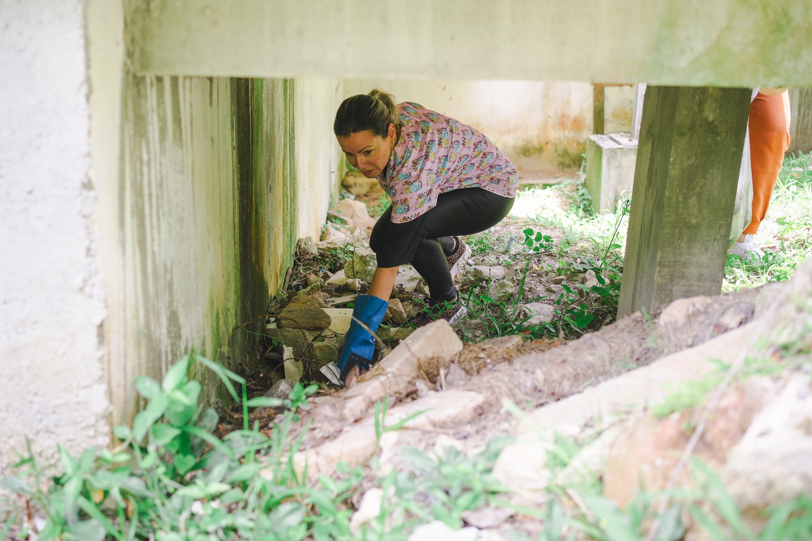 Mulher usando luvas de borracha azuis se abaixa para recolher entulhos e galhos embaixo de uma estrutura elevada de concreto, próxima a uma parede com manchas de umidade e mofo. O local tem vegetação rasteira e pedaços de pedras espalhados pelo chão.