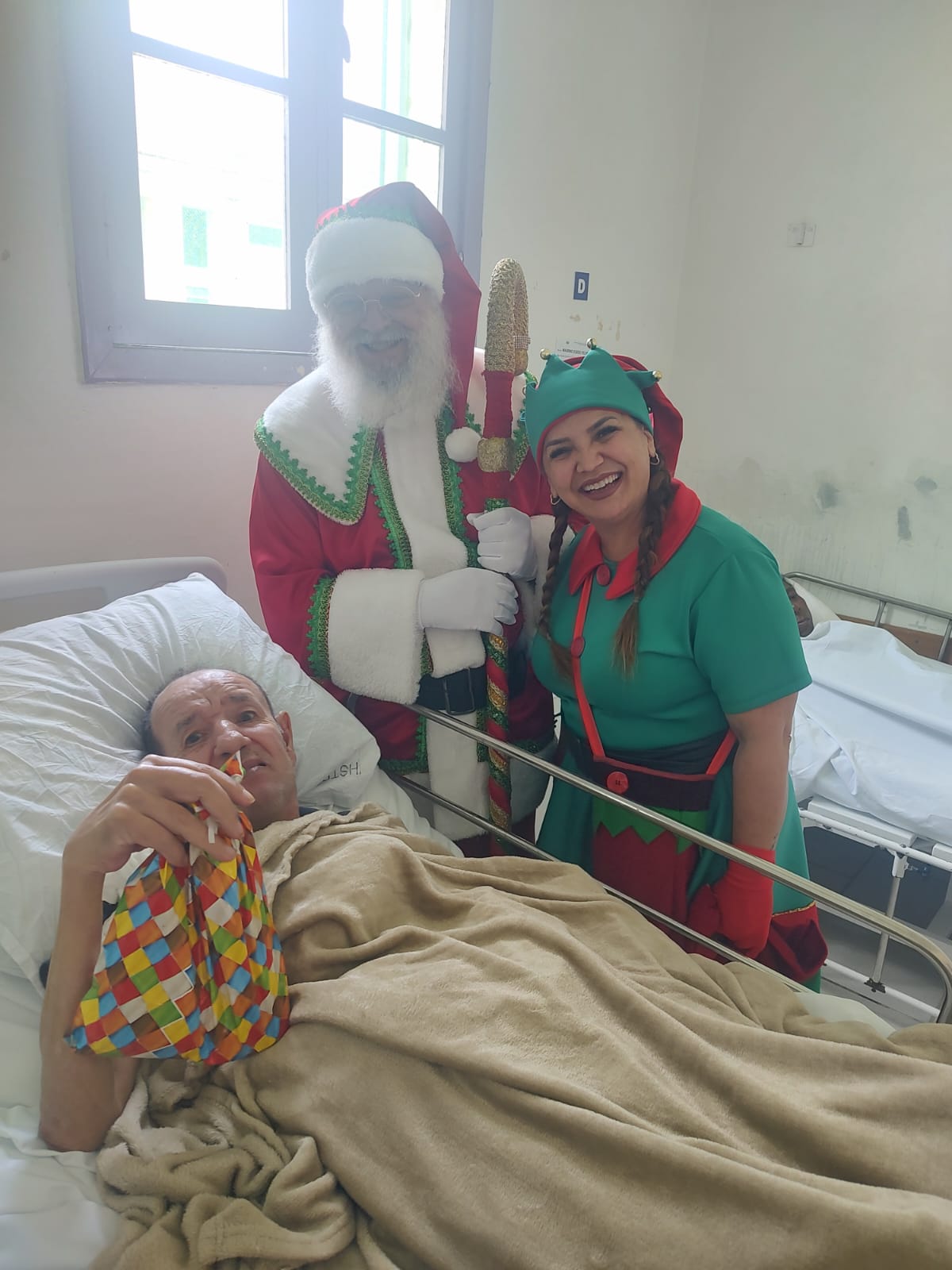 A imagem mostra um momento de visita natalina a um paciente hospitalizado.  No leito, um homem está deitado, coberto com um cobertor bege, segurando um presente embrulhado em papel colorido. Ao lado dele, estão duas pessoas fantasiadas: uma como Papai Noel, com roupas vermelhas e detalhes verdes e dourados, e outra como ajudante/duende de Natal, vestida de verde com detalhes vermelhos e dourados.  Ambos sorriem calorosamente para a câmera, transmitindo um clima de alegria e acolhimento, enquanto o quarto hospitalar é iluminado pela luz natural que entra pela janela ao fundo.