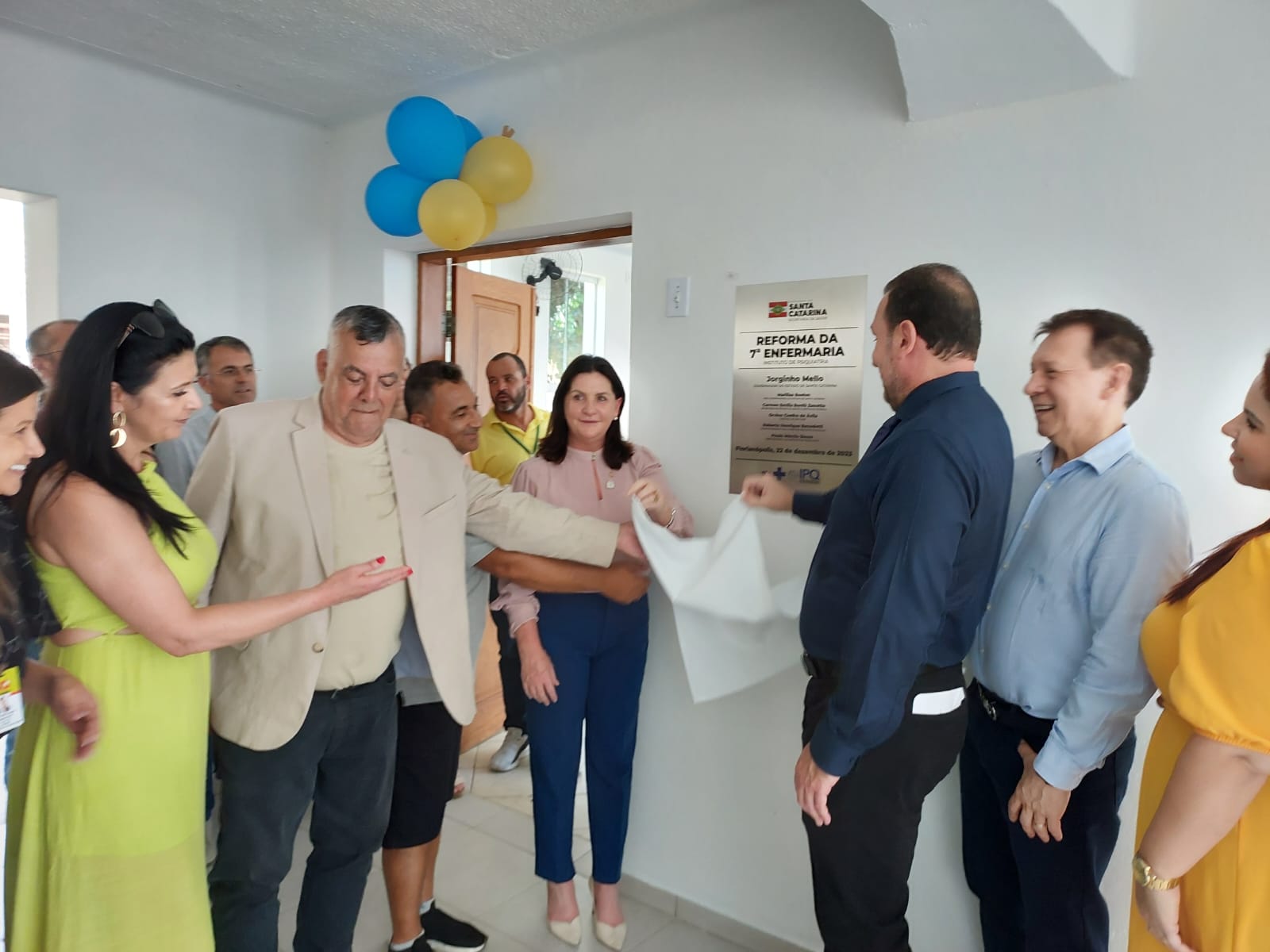 A imagem mostra um grupo de pessoas participando de uma cerimônia de inauguração ou entrega de obra. No centro, dois homens puxam um pano branco que cobria uma placa comemorativa na parede, que anuncia a "Reforma da 7ª Enfermaria" com o nome de Jorginho Mello, governador de Santa Catarina. Há balões azuis e amarelos na parte superior da porta ao fundo, e as pessoas ao redor parecem sorrir e interagir de forma descontraída. O ambiente é interno, com paredes brancas e piso claro.