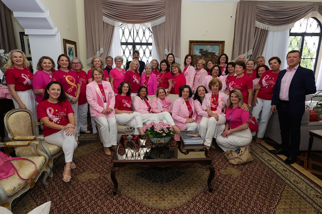 Grupo grande de mulheres e alguns homens reunidos em uma sala elegante, com cortinas e móveis clássicos. A maioria usa camisetas rosa ou vermelhas com o laço símbolo do Outubro Rosa, em alusão à campanha de prevenção ao câncer de mama. Algumas pessoas estão sentadas e outras em pé, posando para a foto com sorrisos. Ao centro da imagem, há uma mesa de centro com arranjo de flores e revistas. Ambiente iluminado por luz natural.