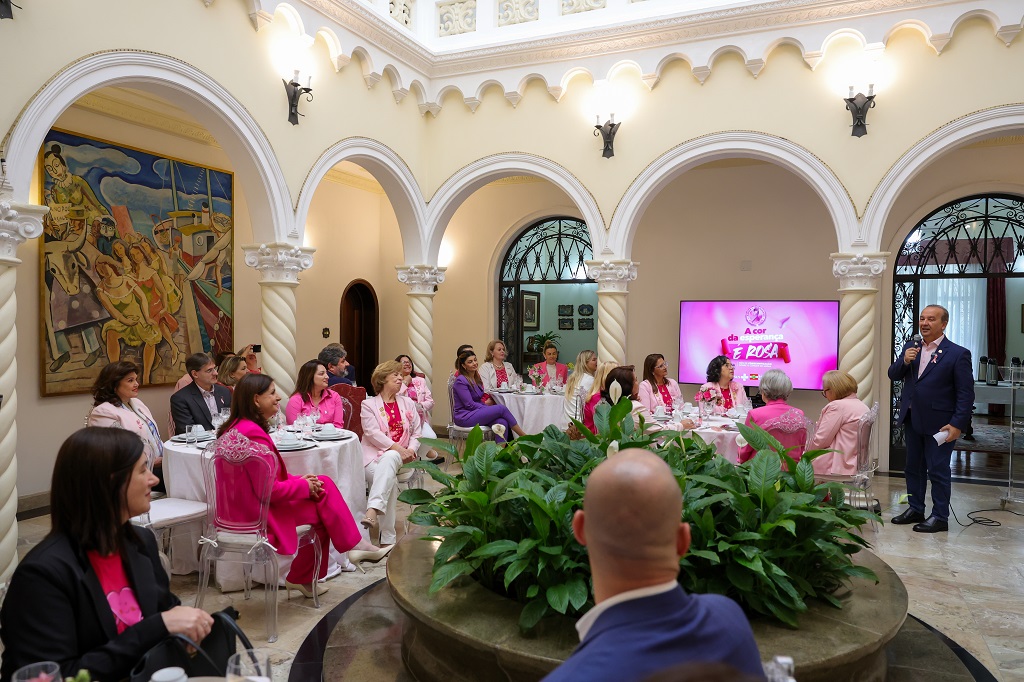 Foto de um evento formal em um salão com arquitetura clássica, colunas brancas em espiral e uma grande planta decorativa ao centro. Várias pessoas, majoritariamente mulheres, estão sentadas ao redor de mesas redondas, vestindo tons de rosa e branco, em alusão à campanha Outubro Rosa. Um homem em pé, também com um laço rosa na lapela, discursa ao microfone próximo a uma tela que exibe o slogan “da cor que é rosa”. Ao fundo, vê-se uma grande pintura colorida com temática marítima.