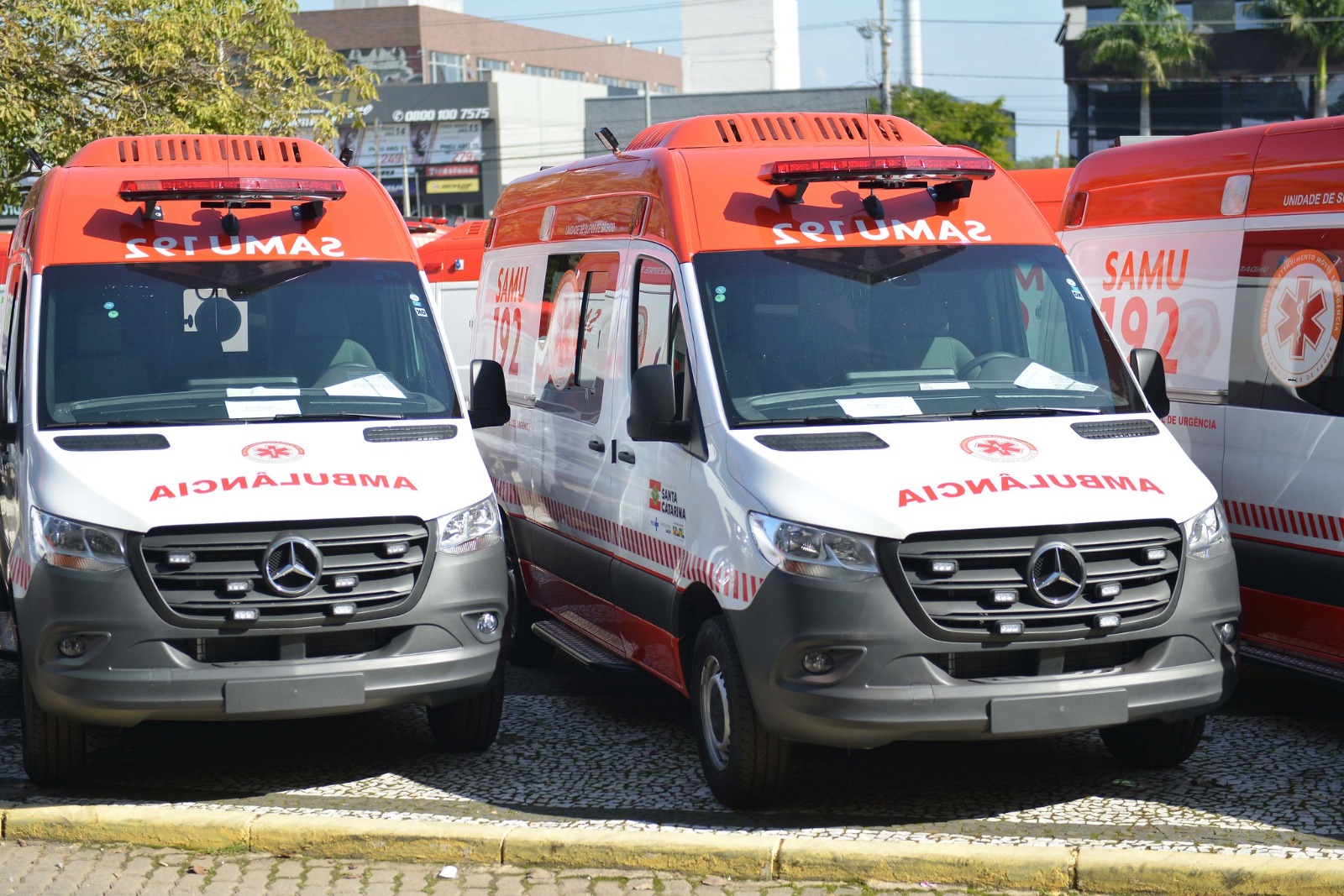 A imagem mostra duas ambulâncias do SAMU (Serviço de Atendimento Móvel de Urgência) estacionadas lado a lado, ambas da marca Mercedes-Benz, com pintura branca e detalhes em vermelho. No capô e na parte frontal superior está escrito “AMBULÂNCIA” e “SAMU 192” (número de emergência). Nas laterais, há o logotipo do SAMU e a bandeira de Santa Catarina, indicando atuação no estado. Ao fundo, outras ambulâncias semelhantes estão estacionadas, e é possível ver parte de uma área urbana com prédios, árvores e placas comerciais.