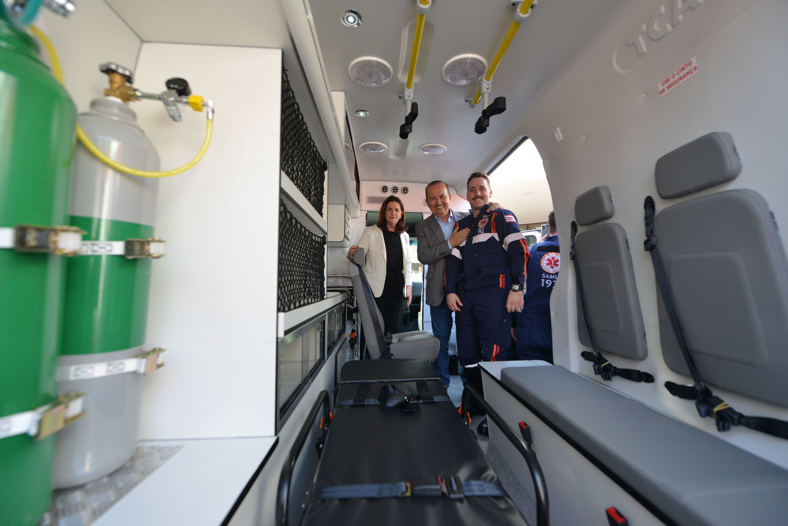 Interior de uma ambulância equipada, com cilindros de oxigênio, maca e assentos. Ao fundo, três pessoas estão em pé, sorrindo: uma mulher de blazer claro, um homem de terno cinza e um profissional do SAMU com uniforme azul escuro com detalhes vermelhos. Ao lado, parcialmente visível, outro profissional do SAMU.