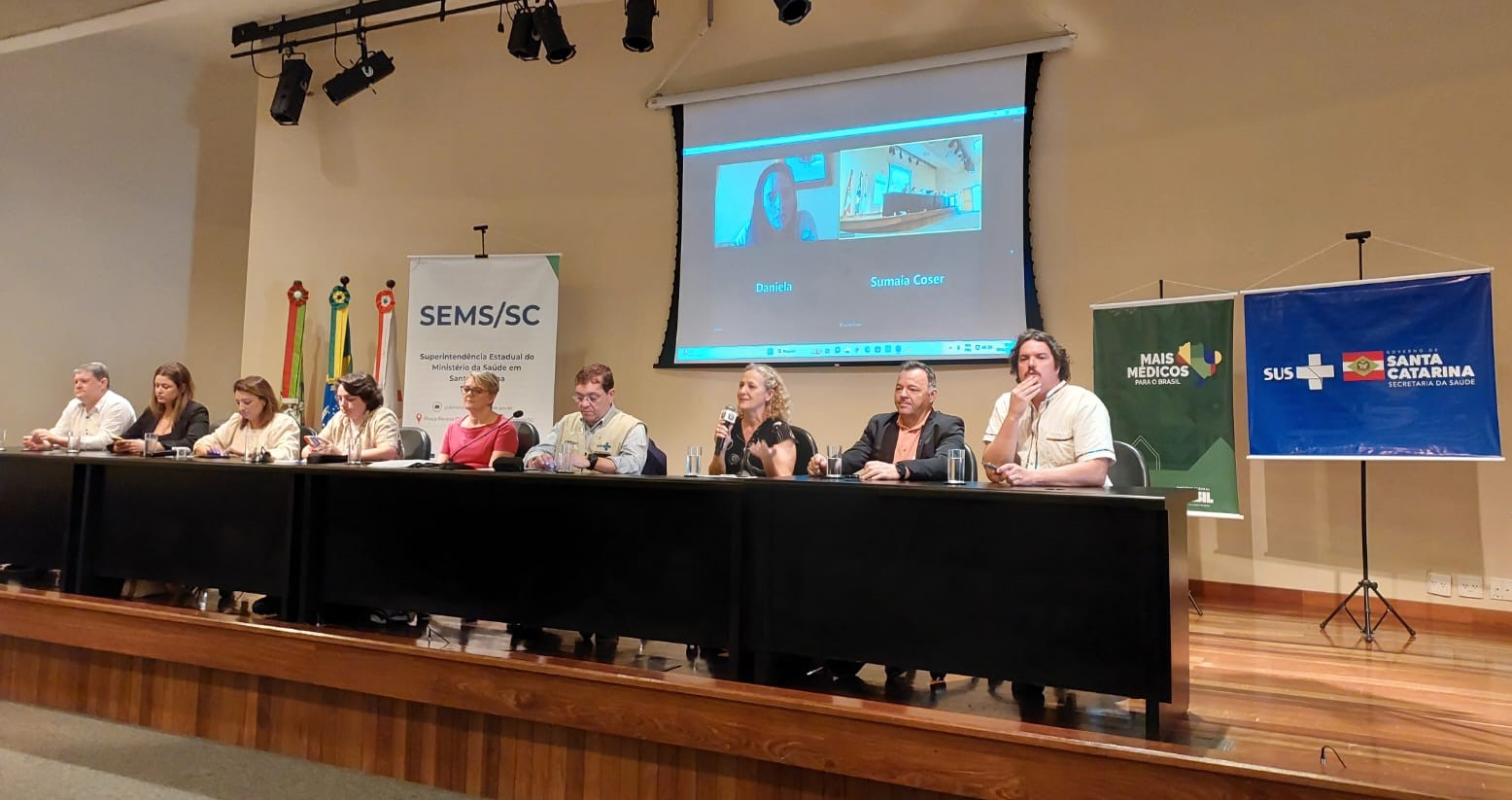 A imagem mostra um evento oficial em um auditório, com nove pessoas sentadas lado a lado em uma longa mesa no palco. No centro, uma mulher fala ao microfone. Ao fundo, há uma projeção de videoconferência com duas participantes remotas (“Daniela” e “Sumaia Coser”).  Também estão presentes banners e faixas institucionais:  SEMS/SC — Superintendência Estadual do Ministério da Saúde em Santa Catarina.  Mais Médicos para o Brasil.  SUS + Governo de Santa Catarina — Secretaria de Estado da Saúde.  Atrás do banner da SEMS/SC, aparecem as bandeiras do Brasil e de Santa Catarina. O ambiente tem iluminação direcionada e piso de madeira, típico de auditórios de eventos governamentais.