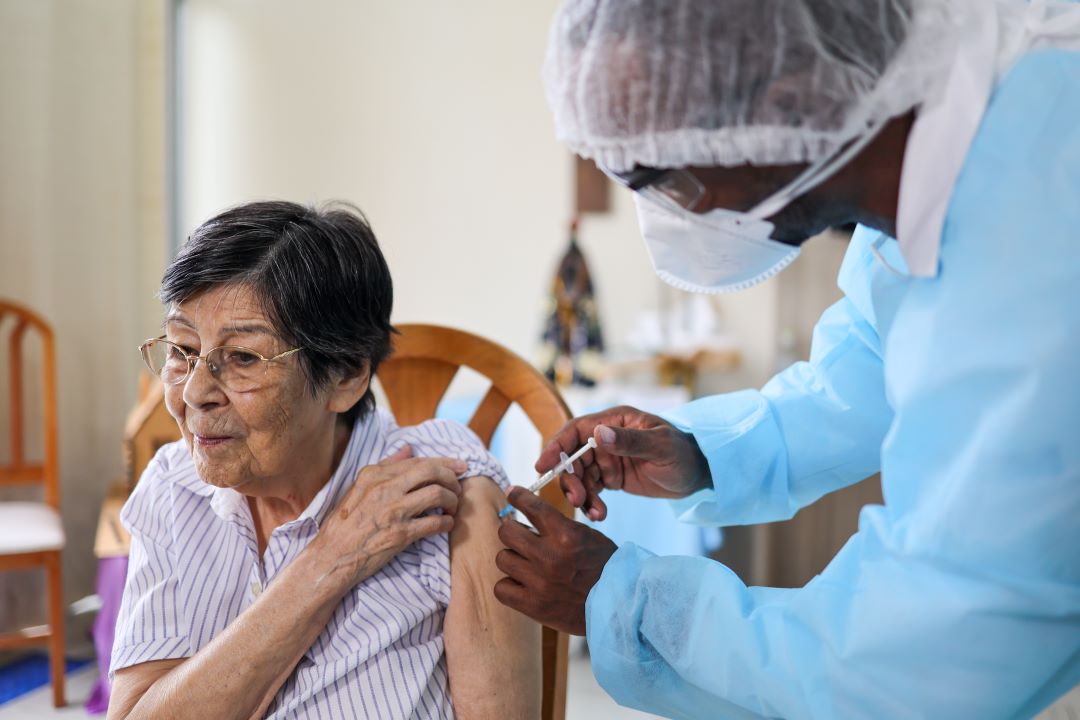 Profissional de saúde com máscara e avental aplica vacina no braço de uma idosa de óculos, sentada em uma cadeira, durante campanha de imunização.