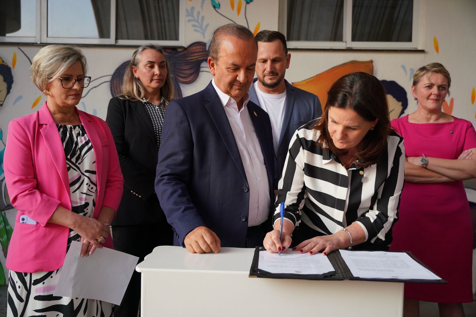 A imagem mostra seis pessoas, quatro mulheres e dois homens, participando de um ato formal de assinatura. No centro, uma mulher de cabelos castanhos, vestindo camisa listrada em preto e branco, assina um documento sobre uma mesa branca, usando uma caneta azul. Ao lado dela, um homem de terno azul marinho e camisa clara observa o ato, enquanto as demais pessoas assistem ao fundo. O cenário é interno, com parede pintada com ilustrações coloridas de crianças, sugerindo um ambiente ligado ao público infantil, possivelmente um hospital ou instituição de acolhimento.