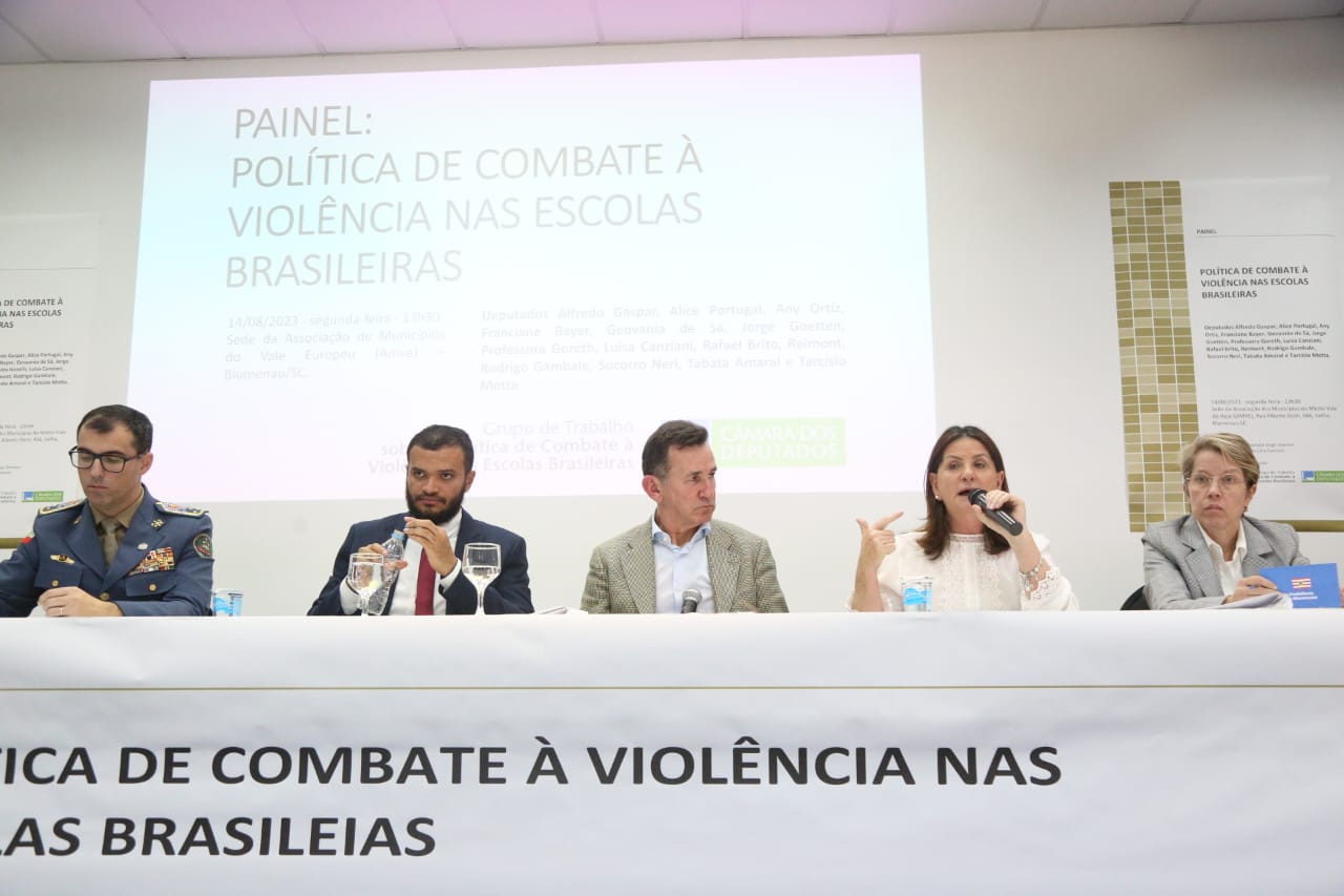 Cinco pessoas sentadas à mesa em audiência pública sobre a política de combate à violência nas escolas brasileiras, com painel projetado ao fundo e faixa com o título do evento na frente da mesa.