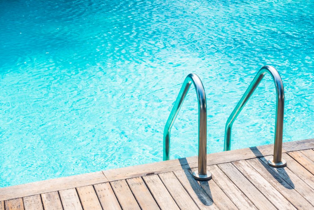 A imagem mostra uma piscina com água cristalina de tom azul claro, refletindo a luz do sol. No canto direito, há uma escada de metal prateado que desce para a piscina, fixada sobre um deque de madeira clara. A cena transmite uma sensação de tranquilidade e convida para um mergulho refrescante em um dia ensolarado.