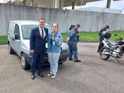 Homem de terno e mulher de roupa casual posam sorrindo em frente a um veículo utilitário prata, com chave na mão, enquanto ao fundo bombeiros e policiais estão próximos a uma motocicleta.