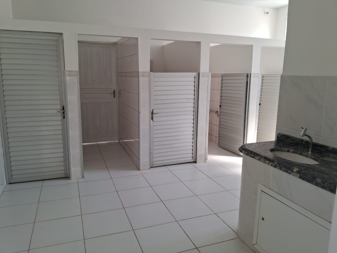 A imagem mostra um banheiro coletivo com várias cabines individuais, cada uma com porta de veneziana branca e paredes revestidas com azulejos claros. No lado direito, há uma pia com bancada de granito escuro e cuba embutida, instalada sobre um gabinete branco. O piso é de cerâmica branca com rejunte claro, e o ambiente está bem iluminado pela luz natural que entra por uma abertura na parte superior da parede. Tudo parece limpo, organizado e em bom estado de conservação.