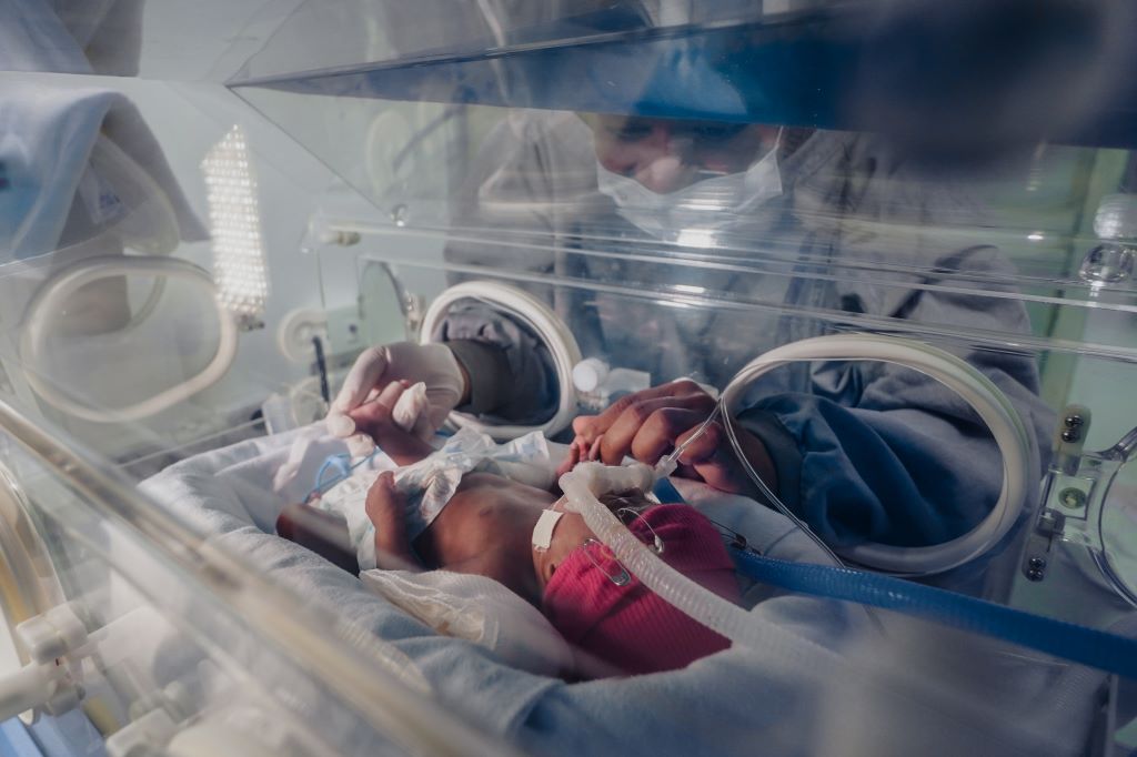 Recém-nascido prematuro deitado em uma incubadora hospitalar, usando gorro rosa e conectado a tubos de respiração e monitoramento. Um profissional de saúde, vestindo máscara cirúrgica e avental, segura delicadamente as mãos do bebê através das aberturas da incubadora, transmitindo cuidado e atenção.