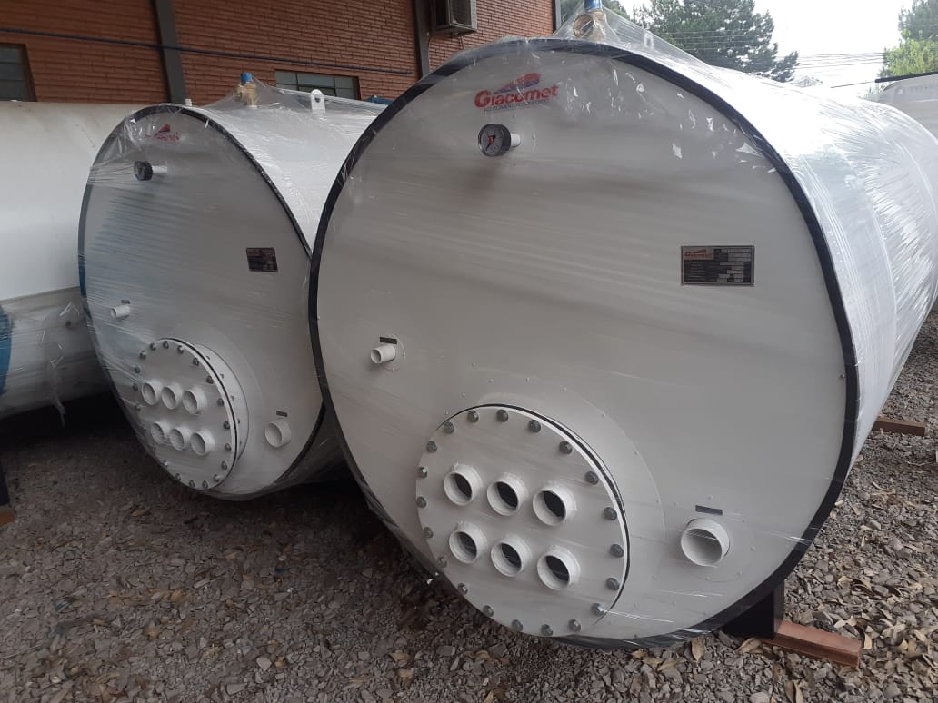 Dois grandes boilers cilíndricos brancos, embalados com filme plástico, posicionados lado a lado em área externa com piso de cascalho.