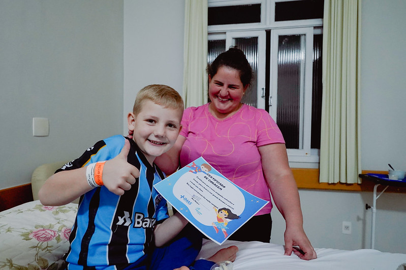 Menino loiro com camiseta do Grêmio sorri e faz sinal de positivo enquanto segura um certificado de "Super-Herói da Coragem". Ele está sentado em uma cama, ao lado de uma mulher, possivelmente sua mãe, que também sorri. Ambos parecem estar em um quarto hospitalar.
