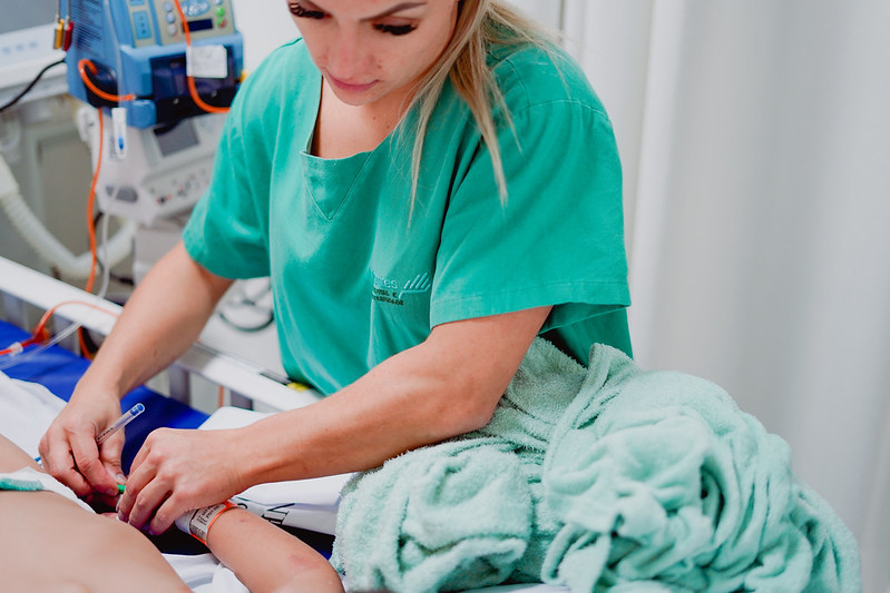 Profissional de saúde loira, vestindo roupa hospitalar verde, realiza procedimento no braço de um paciente deitado, inserindo ou ajustando um acesso venoso. Ao lado, há toalhas verdes enroladas sobre a cama hospitalar e equipamentos médicos ao fundo.