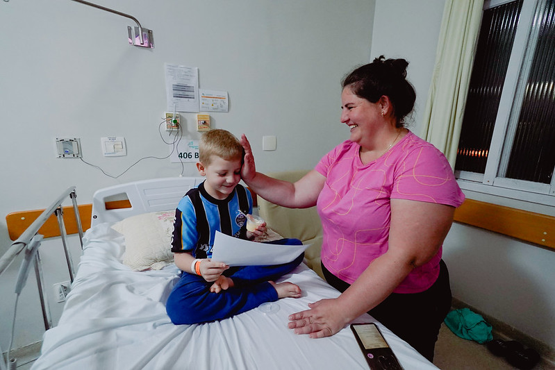 Menino loiro sentado em uma cama hospitalar, vestindo camiseta do Grêmio, sorri enquanto segura um certificado e um pirulito. Ao seu lado, uma mulher sorridente com blusa rosa, provavelmente sua mãe, o parabeniza com um toque carinhoso na cabeça. Ambos parecem felizes em um quarto de hospital.