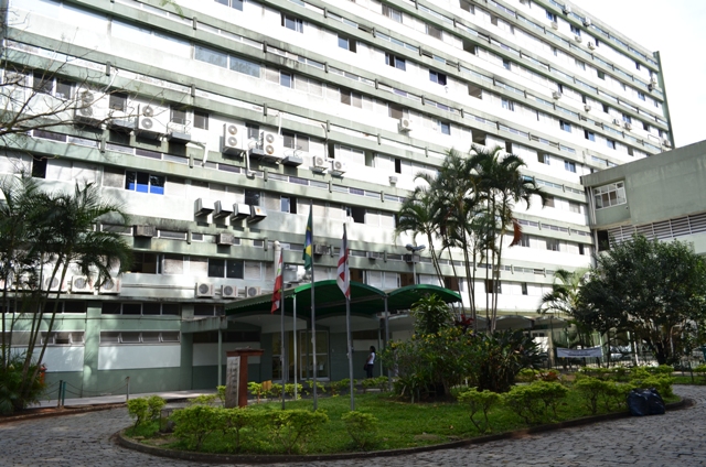 Vista externa de um prédio hospitalar de vários andares, com fachada esverdeada e fileiras de aparelhos de ar-condicionado. Na entrada, há bandeiras hasteadas e uma área ajardinada com coqueiros e arbustos. O local está bem iluminado pela luz natural e apresenta aspecto institucional.